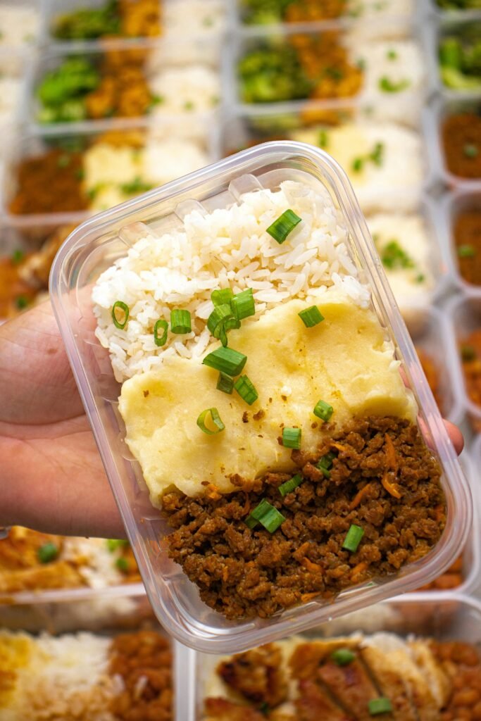 pexels-photo-30635710-30635710 Close-up of a hand holding a meal prep container with rice, mashed potatoes, and ground meat garnished with green onions.