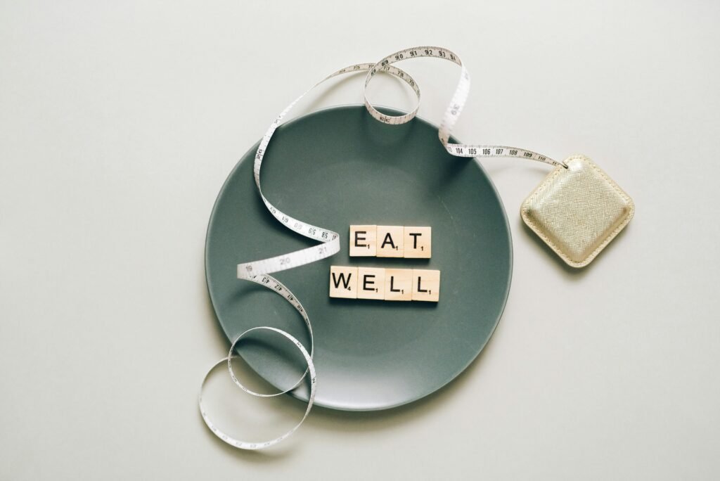 pexels-photo-5842230-5842230 Flat lay of scrabble tiles spelling 'eat well' on a plate with a tape measure, promoting healthy eating.