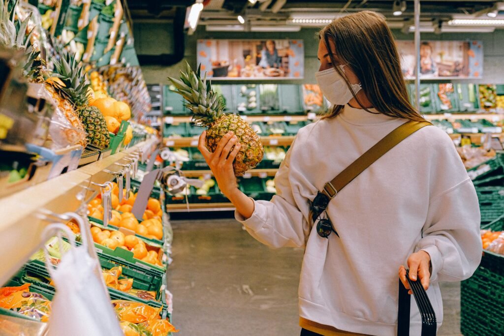 Woman in mask chooses pineapple in a supermarket for safe shopping during pandemic.